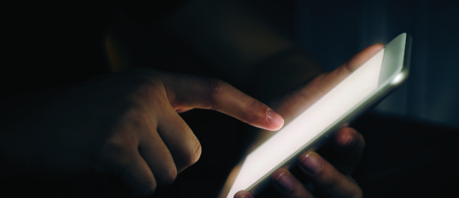 A hand holds a mobile device in the darkness. Light is cast across the hand as they check updates.